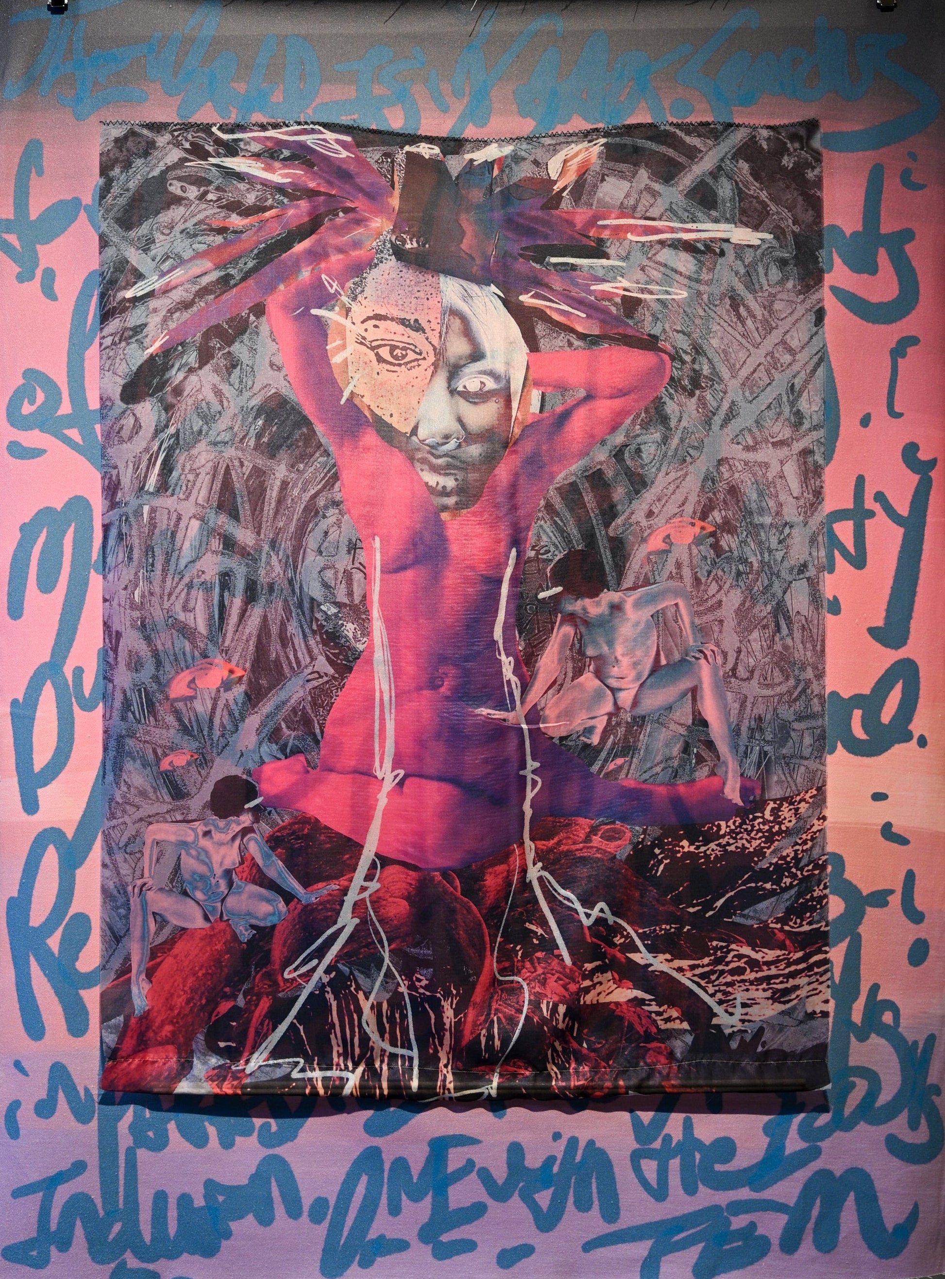 A large textile wall hanging or tapestry displayed on a gallery wall, featuring an abstract central figure in red and black with blue graffiti-style lettering around the border.